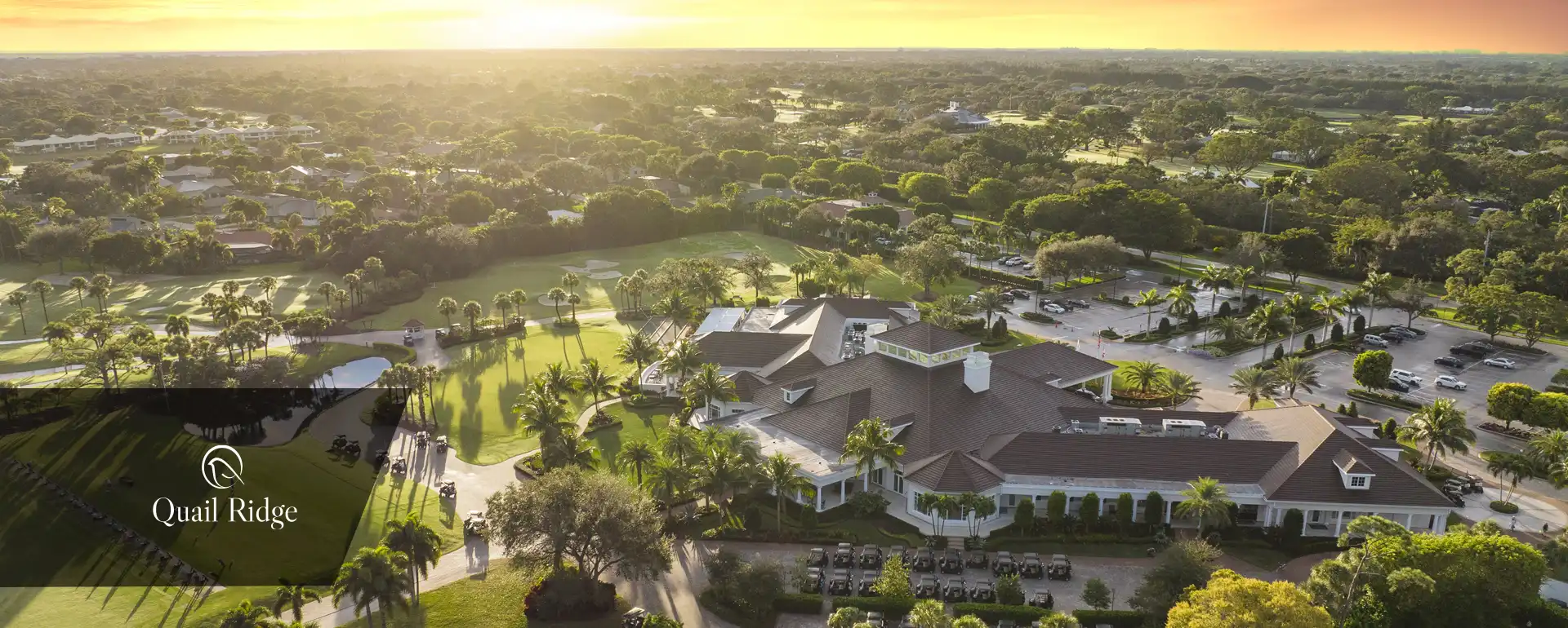 Aerial view of golf clubhouse and course at sunrise with palm trees and surrounding homes.