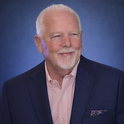 Older man with white hair and goatee in suit smiling against blue background