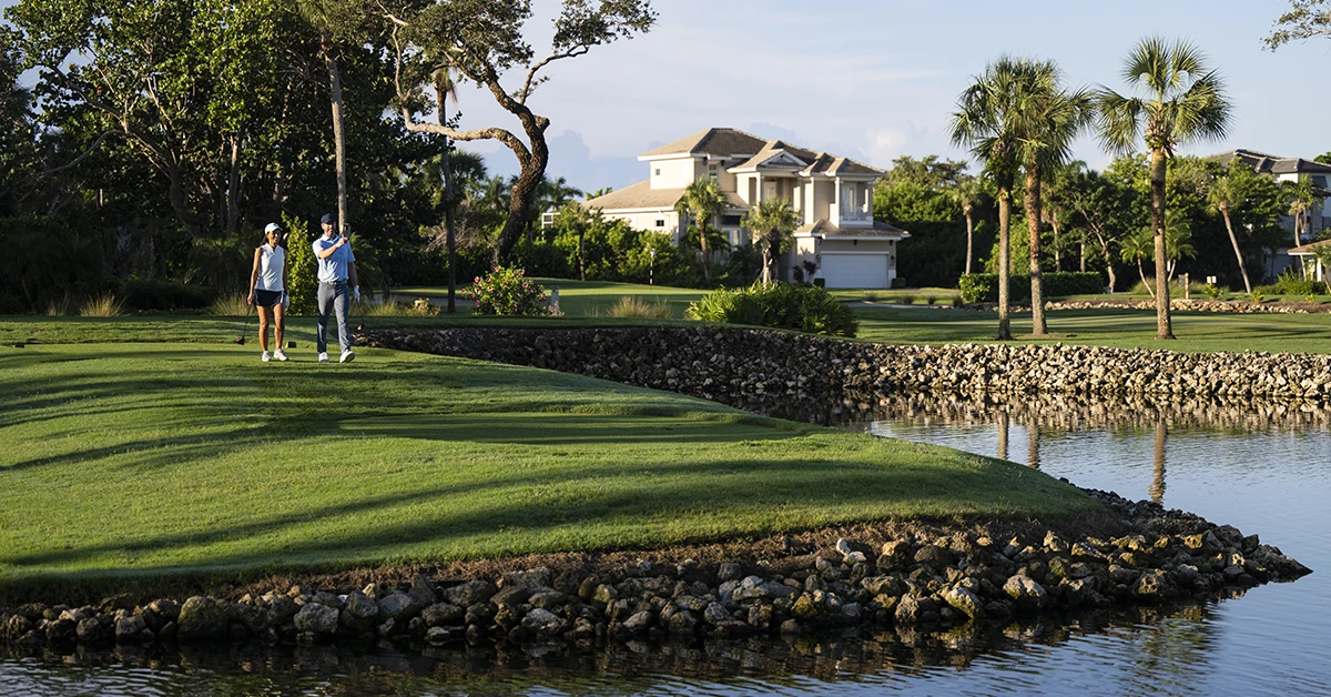 Two golfers walking on fairway beside water near palm trees and home